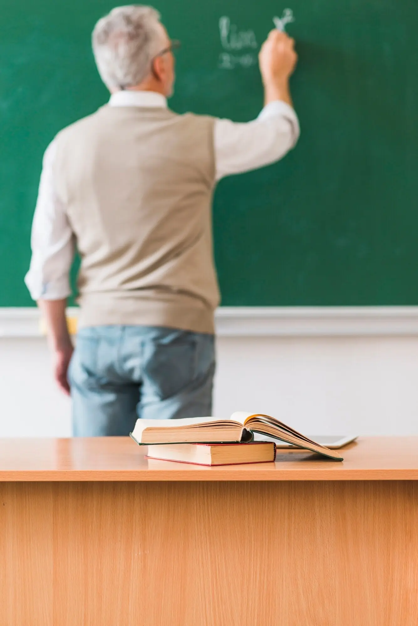 Mathematiklehrer schreibt an die Kreidetafel neben einem Tisch mit Büchern.